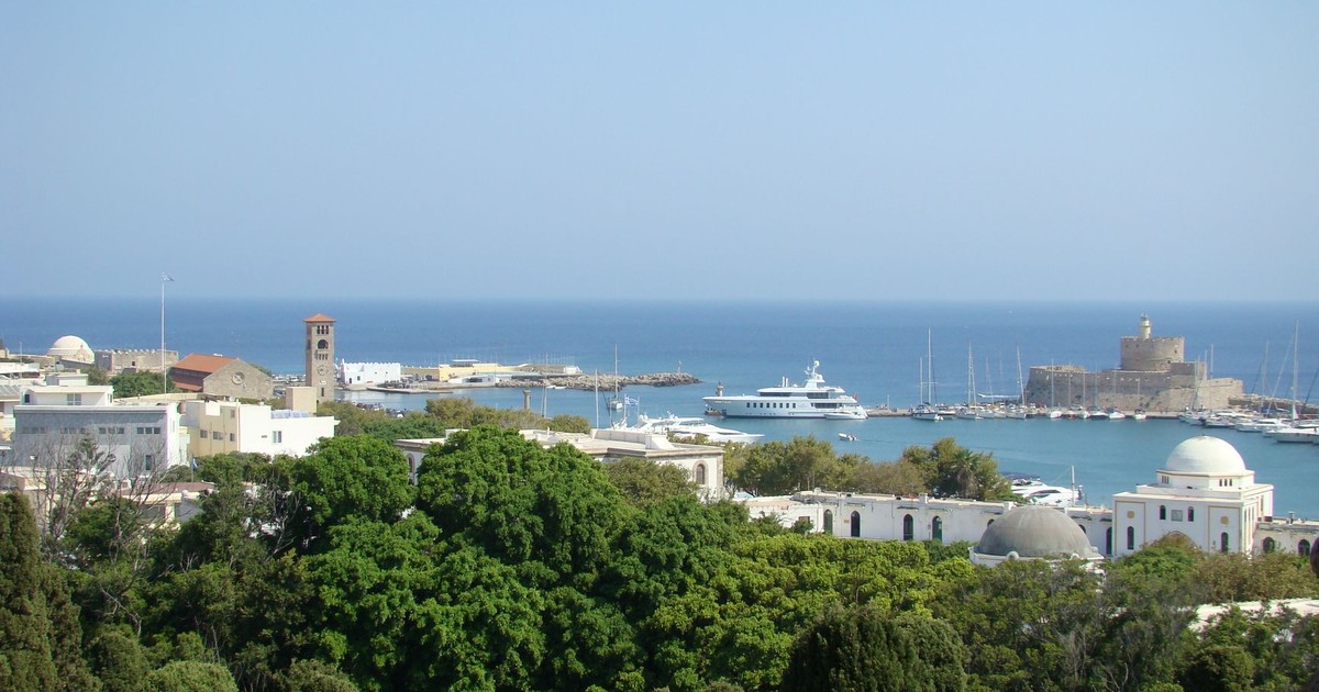 A row of white motor yachts and sailing boats berthed at Mandraki Harbour in Rhodes Town, with the medieval clock tower and fort visible in the background
